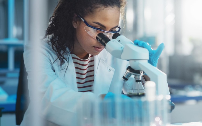 women in lab coat looks through microscope