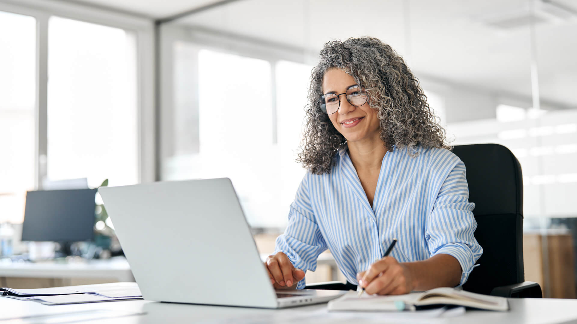 woman smiling and sitting at a desk on her laptop and notebook